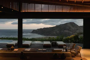 Interior view of a luxury villa's open living space with wooden ceiling slats, modern sofas and armchairs in the foreground, leading to an infinity pool deck with teak loungers and a sweeping view of the ocean, mountains, and dramatic cloudy sky at dusk.
