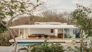 Modern tropical villa with white concrete cantilevered roof, open living room with grey sofas, wooden dining area, and turquoise swimming pool surrounded by lush tropical trees.