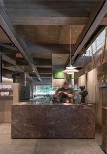 Modern industrial-style coffee shop interior featuring a large dark marble service counter, concrete ceiling, wooden walls, and a barista working behind the counter with coffee equipment.