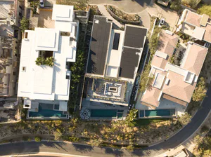 Aerial photograph of a modern luxury villa with white concrete architecture, dark solar panel roofs, rectangular swimming pools, and landscaped courtyards in a desert residential area.