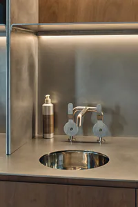 Close-up of a round stainless steel undermount sink with a unique industrial-style metal faucet featuring circular handles, set into a seamless stainless steel countertop with warm wood cabinetry below and under-cabinet lighting.