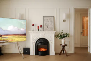 Modern classic living room with ornate white fireplace mantel, black electric fireplace insert, large landscape painting TV on a white stand, red candles, framed heart artwork, and a side table with white flower vase.