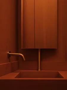 Modern bathroom vanity featuring a square concrete sink, a tall vertical faucet, and a wall-mounted curved spout, all set against warm reddish-brown wood paneling and cabinetry.