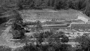 森林山坡大型工地鳥瞰圖，可見多層石材擋土牆、挖土機、土路與建築基礎。