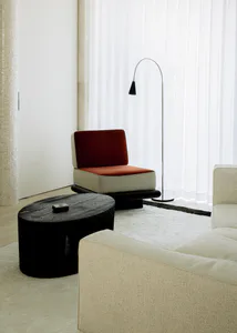 Modern minimalist living room corner featuring a red velvet and beige armchair with black base, a round black wood coffee table, a tall arc floor lamp, and a cream bouclé sofa on a white rug.