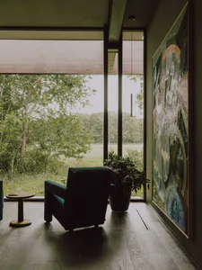 Modern living room interior featuring a teal leather armchair, round wooden side table, large abstract painting, potted plant, and expansive floor-to-ceiling windows overlooking a green forest and meadow.