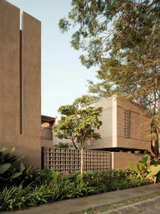 Modern house with exposed concrete walls, brick detailing, and a lush tropical garden featuring various ferns and trees under a clear sky.
