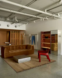 Industrial-style retail showroom with a brown tufted velvet sofa, metallic coffee table, red plastic chair, woven jute rug, and exposed white ceiling pipes.