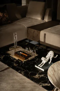 Close-up of a luxurious living room coffee table with black marble top, featuring a white ceramic leopard sculpture on a book, a wooden cigar ashtray with a cigar, two whiskey glasses, and a tall glass decanter, with a neutral modern sectional sofa in the background.