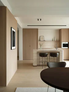 Modern minimalist kitchen with a large travertine stone island, two black leather bar stools, light oak wood cabinetry, and a round dark wood dining table in the foreground.