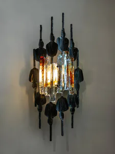 A sculptural wall sconce mounted on a light gray wall, featuring dark forged iron figures surrounding a central cluster of illuminated, textured glass panels in amber, clear, and red tones.