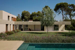 Modern minimalist concrete house with large glass openings, white curtains, and a lush garden with purple flowers leading to a turquoise swimming pool under a clear blue sky.