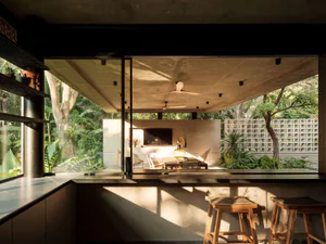 Modern tropical indoor-outdoor living space featuring a concrete bar counter with wooden stools, large glass openings, and a lush garden courtyard with daybeds and ceiling fans.