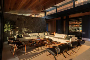 Modern open-plan living room with large L-shaped sectional sofa, wooden coffee tables, black leather and wood sling chairs, a prominent stone feature wall, wooden ceiling, and a built-in bar area in the background.