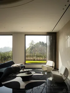 Modern minimalist living room featuring two white sculptural lounge chairs, a dark curved sofa with fur throw, black and white circular rug, and large floor-to-ceiling windows overlooking a green garden and trees.