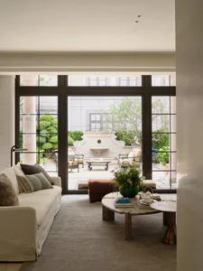 Modern living room with off-white sofa, travertine coffee table, and large glass doors opening to a manicured courtyard with a stone fountain.