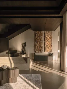 Modern minimalist entryway featuring travertine stairs, fluted stone furniture, two large organic stone wall sculptures, and a dark wood sculpture under dramatic natural light.