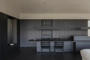Modern minimalist kitchen featuring a long dark wood dining island with two black chairs, matching dark wood cabinetry, open shelving with ceramics, and a concrete ceiling with pendant lights.