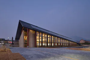 Exterior view of a long modern public library building at dusk, featuring a dramatic pitched roof, extensive floor-to-ceiling glass walls revealing glowing bookshelves inside, concrete and wood structural elements, set against mountains in Taiwan.