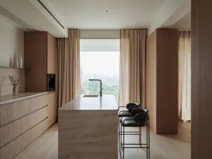 Modern minimalist kitchen with a large travertine stone island, three black leather bar stools, light oak cabinetry, and a large window overlooking green trees.