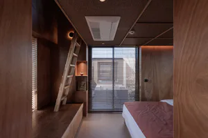 現代小型臥室，滿室木質板材、內建閣樓梯、大型百葉窗以及鋪有赤陶色床單的床鋪。