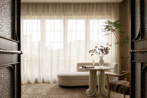 View into a bright, minimalist living room with a curved white chaise, travertine pedestal coffee table, wooden armchair with zebra-print cushion, and large sheer-curtained windows overlooking a balcony.