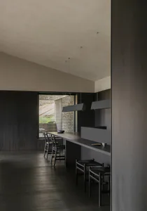 Modern minimalist kitchen featuring a long dark wood island, woven leather bar stools, rectangular pendant lights, and large windows overlooking a stone wall courtyard.
