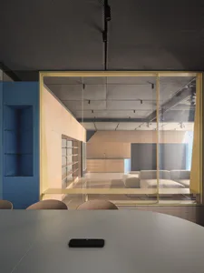 View from a conference table looking through a golden-framed glass partition into a modern open-plan office lounge with white modular sofa and wooden cabinetry.