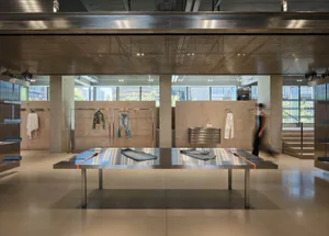 Spacious industrial-style fashion boutique interior featuring raw concrete walls, polished concrete floors, a large stainless steel display table with orange accents, and clothing hanging on metal racks under a perforated metal ceiling.