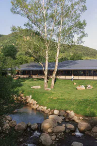 Modern single-story house with large windows situated on a green lawn next to a rocky stream, surrounded by tall birch trees and forested mountains in the background.