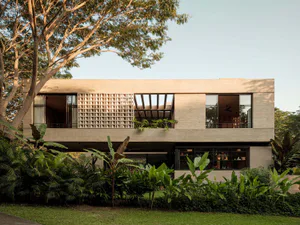 Modern two-story tropical house with exposed concrete walls, large glass windows, and decorative breeze block facade surrounded by lush tropical garden.