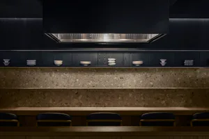 Modern dark kitchen featuring a long terrazzo countertop, light wood dining counter with navy blue chairs, open floating shelves displaying white ceramics, and dramatic under-cabinet lighting.