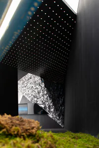 View of a modern interior featuring a large crinkled silver foil sculpture against black walls, a starry LED dotted ceiling, and moss-covered foreground.