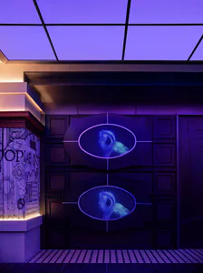 Futuristic digital art installation featuring two large oval LED screens displaying glowing blue eyes, set within ornate dark purple wall panels under a violet ceiling with dramatic theatrical lighting.