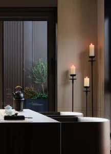 A modern minimalist Japanese-style tea room featuring a dark wood table set with a cast iron teapot, white ceramic teacups, and a small incense holder. Three lit white pillar candles in tall black metal candlestands cast a warm glow against a neutral wall, with a view of a small potted tree through a glass door with vertical blinds.