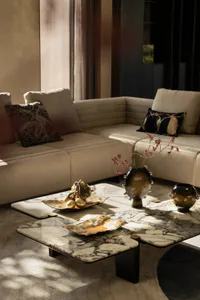 Modern luxury living room with a cream L-shaped sectional sofa, marble coffee tables, gold decorative trays, dark metallic vases with dried branches, and patterned throw pillows.