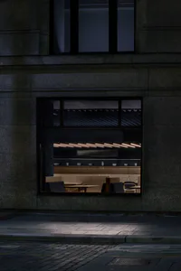 Night view through a large window into a minimalist Japanese restaurant interior featuring a terrazzo counter, wooden dining benches, and linear pendant lighting.