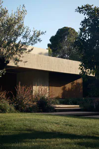 Modern concrete house with extensive wooden slat cladding, surrounded by lush garden with olive trees, pampas grass, and manicured lawn under a clear blue sky.