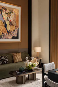 Elegant modern living room interior with a green velvet sofa, patterned throw pillows, a black and gold marble coffee table holding a large floral arrangement and a green ceramic jar, a textured side chair, and a large colorful abstract landscape painting in a gold frame hanging on a warm brown paneled wall.