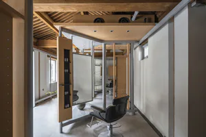 Modern industrial-style hair salon interior featuring exposed wooden ceiling beams, concrete floor, black leather salon chair, and modular wooden storage units with metallic structural columns.