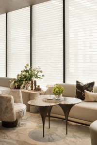 Modern living room featuring a sculptural travertine coffee table, patterned swivel armchair, beige sectional sofa with textured pillows, large floral arrangements, and floor-to-ceiling sheer horizontal blinds.