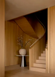 Modern minimalist entryway featuring a curved oak spiral staircase, light wood paneling, and a large stone vase with flowering branches on a conical metal pedestal.
