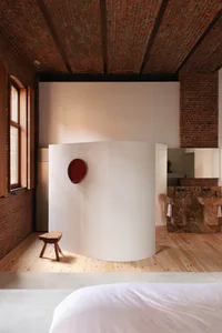 View of a modern loft bathroom featuring a large curved white plaster partition wall with a red circular mirror, exposed red brick walls and ceiling, wooden floor, and marble double vanity in the background, with a wooden stool in the foreground.