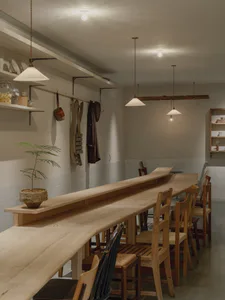 極簡餐廳室內景觀，展示一道極長的弧形實心橡木共享餐桌，搭配木椅、吊燈、開放式層架，以及桌上盆栽植物。