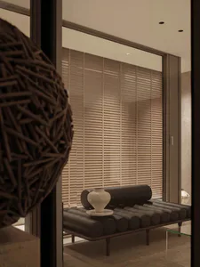 Modern minimalist interior featuring a black leather roller daybed with cylindrical cushions, a white ceramic vase on a marble tray, and floor-to-ceiling light wood venetian blinds, viewed through a dark frame with a woven sculptural object in the foreground.