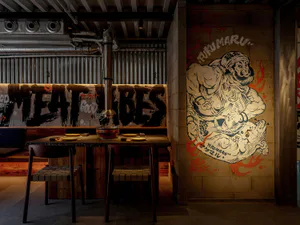 Industrial-style Japanese restaurant dining area featuring a large Fukumaru mural on exposed concrete wall, dark wooden table with hot pot setup, woven leather chairs, and corrugated metal background.