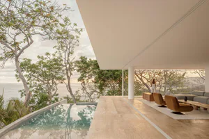 Modern open-air living room with travertine floors, brown velvet swivel chairs, a low wooden coffee table, and a long infinity pool overlooking the ocean and lush trees at sunset.