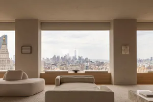 Modern minimalist living room with beige boucle sofas and daybed, large floor-to-ceiling windows revealing the New York City skyline including One World Trade Center and the Woolworth Building under an overcast sky.