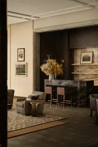 Elegant modern hotel lobby interior with dark marble reception desk, two wooden bar stools, a large dried floral arrangement with bird of paradise flowers, neutral upholstered armchairs on a patterned area rug, and framed artwork on light stone walls.