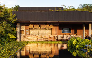 Modern tropical outdoor kitchen and dining pavilion with live edge wood counter, woven pendant lights, stone wall, and reflecting pool surrounded by lush greenery.